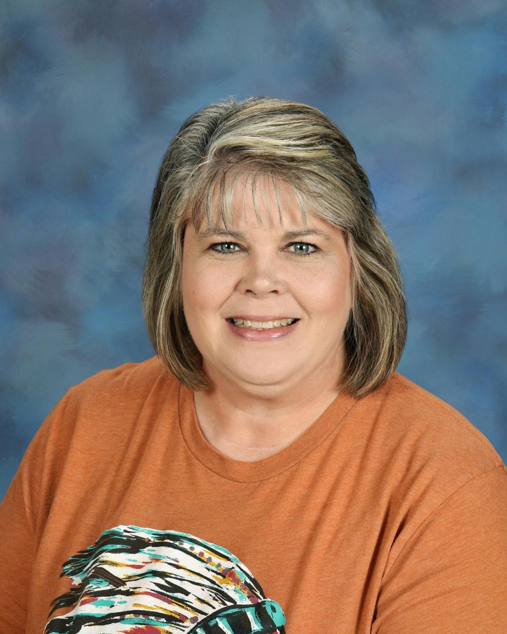 Lady with blond hair, wearing a orange shirt with graphics on the front.  